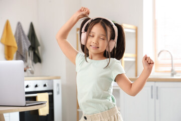 Wall Mural - Little Asian girl with headphones and laptop dancing in kitchen