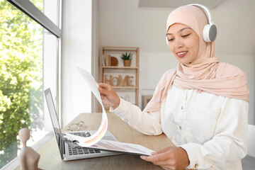 Canvas Print - Young Muslim businesswoman in headphones with clipboard working at home