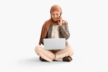 Wall Mural - Young Muslim businesswoman with laptop talking by mobile phone on light background