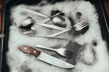 A knife with wooden handle, fork and spoons in the detergent foam on a black oven-tray. Washing dishes concept.