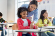 © Home-stock - Teacher helping black girl in classroom, black schoolgirl writing in notebook, sitting at desk with classmates on background