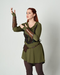 Wall Mural - Close up portrait of beautiful  red haired female model, wearing green and brown medieval fantasy costume with tunic and armour. Standing  with gestural hand poses, isolated on white studio background