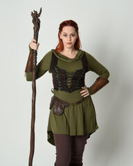 Wall Mural -  portrait of beautiful red haired female model, wearing green medieval fantasy costume with brown tunic armour armour. Standing pose with wood wizard staff weapon, isolated white studio background.