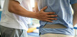 © Sourav Mittal - Close up of a young man with back pain, with his hands on his lower hip and upper waist area while a male physical therapist helps him feel better in the clinic office. The man has bad sciatica in his