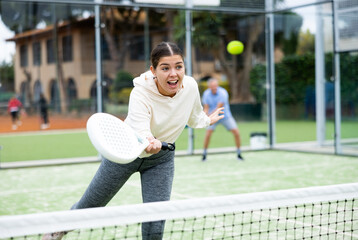 Wall Mural - Emotional young European girl padel player hitting ball with racket on hard court in autumn