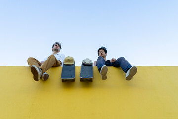 Wall Mural - Two young men with skateboards sitting on a high yellow wall