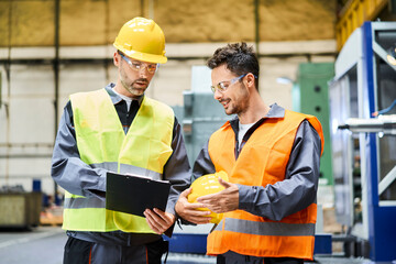 Wall Mural - Two men wearing protective workwear holding clipboard and talking in factory