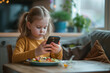 © Patcharaphon - Little girl using mobile phone while having lunch on the table. Screen addiction concept