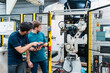 © tunedin - Male coworkers holding digital tablet looking at robotic arm while standing in manufacturing industry