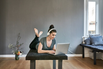 Poster - Young woman with headphones lying on lounge using laptop