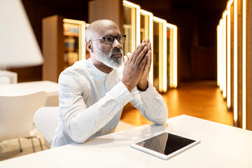 Portrait of pensive mature man sitting at desk in a library with digital tablet