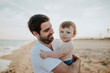 © tunedin - Smiling father carrying baby boy at beach during sunset