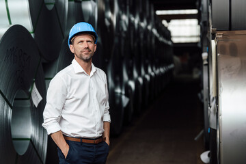 Businessman contemplating with hands in pockets standing by steel rolls in factory