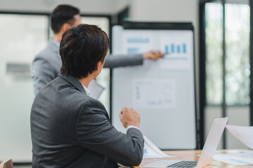 Wall Mural - Businessman listens attentively as colleague presents detailed analysis using charts and graphs on whiteboard during meeting in modern office.