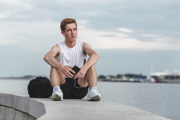 Wall Mural - Young athlete sitting on a wall at riverbank with cell phone and earphones