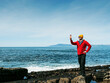 © mark_gusev - A man in red jacket and yellow hat taking picture on smart phone of rough stone coastline. West of Ireland. Travel and tourism. Irish landscape. Ocean in the background.