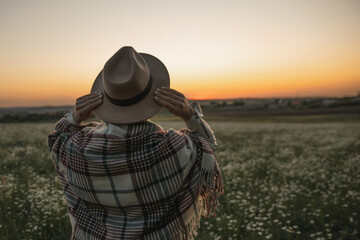 Wall Mural - A woman wearing a hat and a plaid blanket stands in a field of flowers. The sky is orange and the sun is setting.