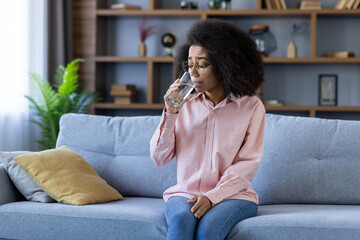 Wall Mural - Young woman sits on a cozy sofa while drinking water in a modern living room setting, showcasing a casual and relaxed moment.