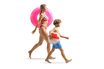 Wall Mural - Full length profile shot of a young woman in bikini carrying a swimming ring and boy with a beach ball