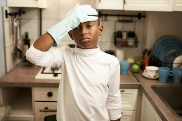 Exhausted upset black teen boy covering forehead with hand in blue gloves, tired of cleaning house and dirty kitchen after baking cake, wearing mockup long-sleeve with copy space for your content