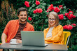 © Masson - man and woman sitting together at outdoor table in a garden setting with roses, surrounded by vibrant red flowers and lush greenery. Both are engaged with a laptop