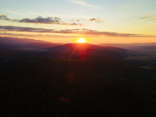 Poster - Sun rays. Green pine forest slopes of mountain range. Bright disk of sun in blue sky slowly sets tops of mountains. Nature. Lens flare in mountains at orange sunset in summer morning aerial view drone