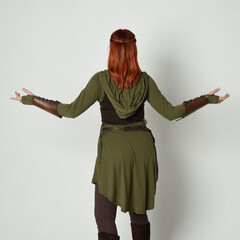 Wall Mural - portrait of beautiful red haired female model, wearing green and brown medieval fantasy costume with tunic and armour. Standing  pose facing away backwards , isolated on white studio background.