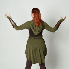 Wall Mural - portrait of beautiful red haired female model, wearing green and brown medieval fantasy costume with tunic and armour. Standing  pose facing away backwards , isolated on white studio background.