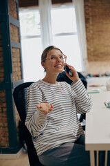 Wall Mural - Young smiling female entrepreneur talking by phone with client while sitting in modern office 