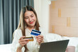 © Ekkasit A Siam - Asian woman smiling while holding credit cards in front of laptop. Long hair, casual outfit. Modern home office setup, beige decor, natural light., pink coffee cup on table, indoor environment.