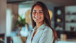 © Alena - Beautiful tanned woman with a soft smile, neat loose hair, and light makeup, wearing a bright shirt, hands in pockets, against a blurred modern office. She exudes success and happiness.
