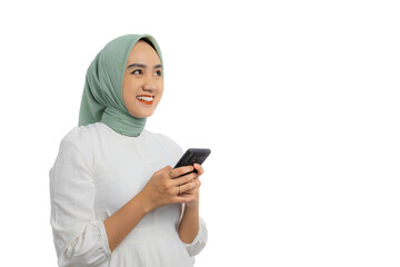 Wall Mural - Happy young Asian woman in green hijab and white blouse holding mobile phone and looking away with friendly smile isolated on white background