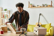 © LIGHTFIELD STUDIOS - A young African American male blogger opens a box in his living room while talking on the phone camera.