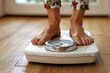 © Sachin - Person standing on a weighing scale, focusing on health and weight management, in a bright home setting with natural light and wooden flooring.