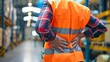 © Irina Ukrainets - Warehouse worker with back pain wearing a safety vest in a busy industrial setting. Focus on workplace safety and health. Ideal for business, industry, and safety related content. AI