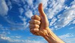 © owen - Close-Up of Hand Giving Thumbs Up Against a Vibrant Blue Sky with White Clouds, Symbolizing Positivity, Success, and Approval in a Bright Outdoor Setting