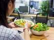 © mirifadapt - Young woman eating a salad in office. Young businesswoman sitting from desk.