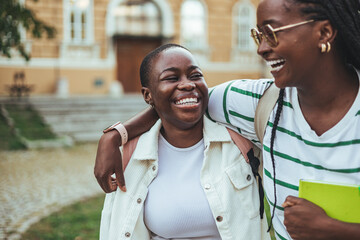 Wall Mural - Two cheerful young black women with backpacks enjoy a break, laughing and sharing a friendly embrace outside their college building.