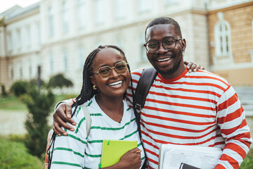 Wall Mural - A joyful black male and female student embrace while holding educational materials, dressed casually on a university campus, radiating enthusiasm for learning.