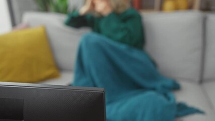 Wall Mural - A young woman yawns while sitting on a couch, covered in a teal blanket in a cozy living room.