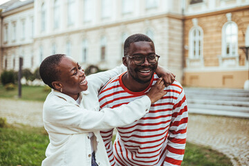 Wall Mural - A smiling black male and female college students share a playful moment outside an academic building, dressed in casual attire suitable for a day of studies and relaxation.