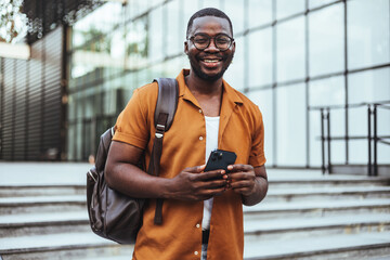 A smiling black male student, wearing casual attire with a backpack, uses a smartphone while standing on university steps.