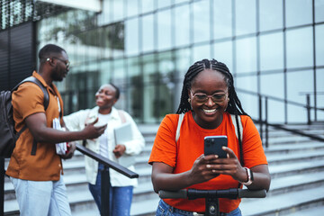 Wall Mural - A radiant black female student in a vibrant orange shirt smiles while using her smartphone, standing near a university building with diverse classmates in the background.