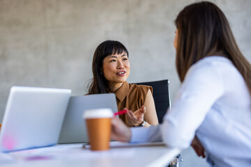 Wall Mural - An Asian and a Caucasian businesswoman discuss intently, laptops open and coffee at hand, in a modern office setting.