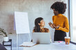 © Dragana Gordic - An Asian businesswoman and a black businesswoman discuss strategies at work, both professionally dressed, using a laptop and notes, in a modern office setting.