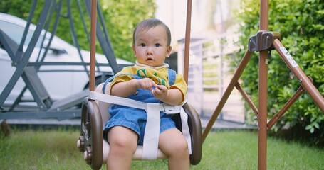 Wall Mural - Happy cute adorable asian baby infant child toddler sitting on a swing with seatbelt in a park enjoy recreation carefree relaxation in summertime at home