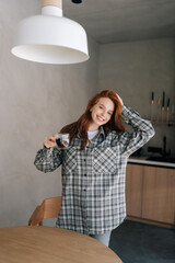 Vertical portrait of cheerful young woman enjoying holding cup drinking coffee standing in kitchen on sunny morning, smiling looking at camera. Happy female relaxing on caffeine break alone.