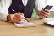 © Studio Romantic - Cropped photo of business man writing tasks and to do list in notepad sitting at the desk with smartphone in hand at home or at office planning daily appointment in plan schedule. Planner concept.