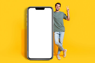 Full length photo of good mood cute guy wear khaki t-shirt showing v-sign modern device empty space isolated yellow color background