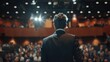 © pitu - A well-dressed businessman stands with his back to the camera, addressing an attentive audience in a large conference hall. He appears confident and poised as he presents his keynote,
